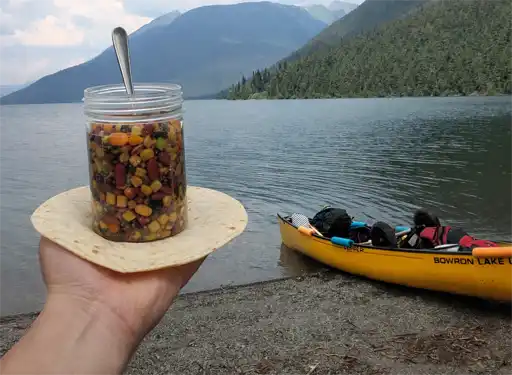 Cold-soak backpacking lunch with tortilla wrap during shoreline canoe trip stop on Bowron Lakes
