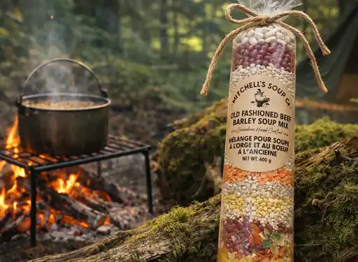 A package of Mitchells Old Fashioned Beef Barley soup leaning on a moss filled tree root with a campsite background and a pot of soup on a campfire
