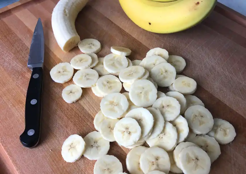 Showing whole bananas and sliced bananas with a paring knife on a wooden cutting board
