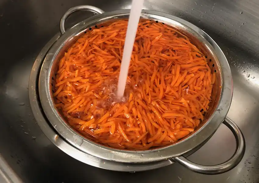 A photo showing shredded carrots being run under cold water to stop the cooking process
