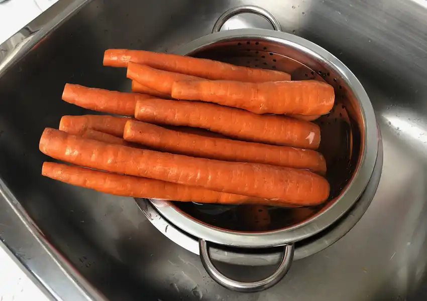 Showing a bowl and colander with whole carrots being washed in a sink