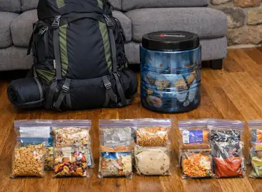 Showing a backpack, bear can, and 4 days of packaged meals on a living room floor